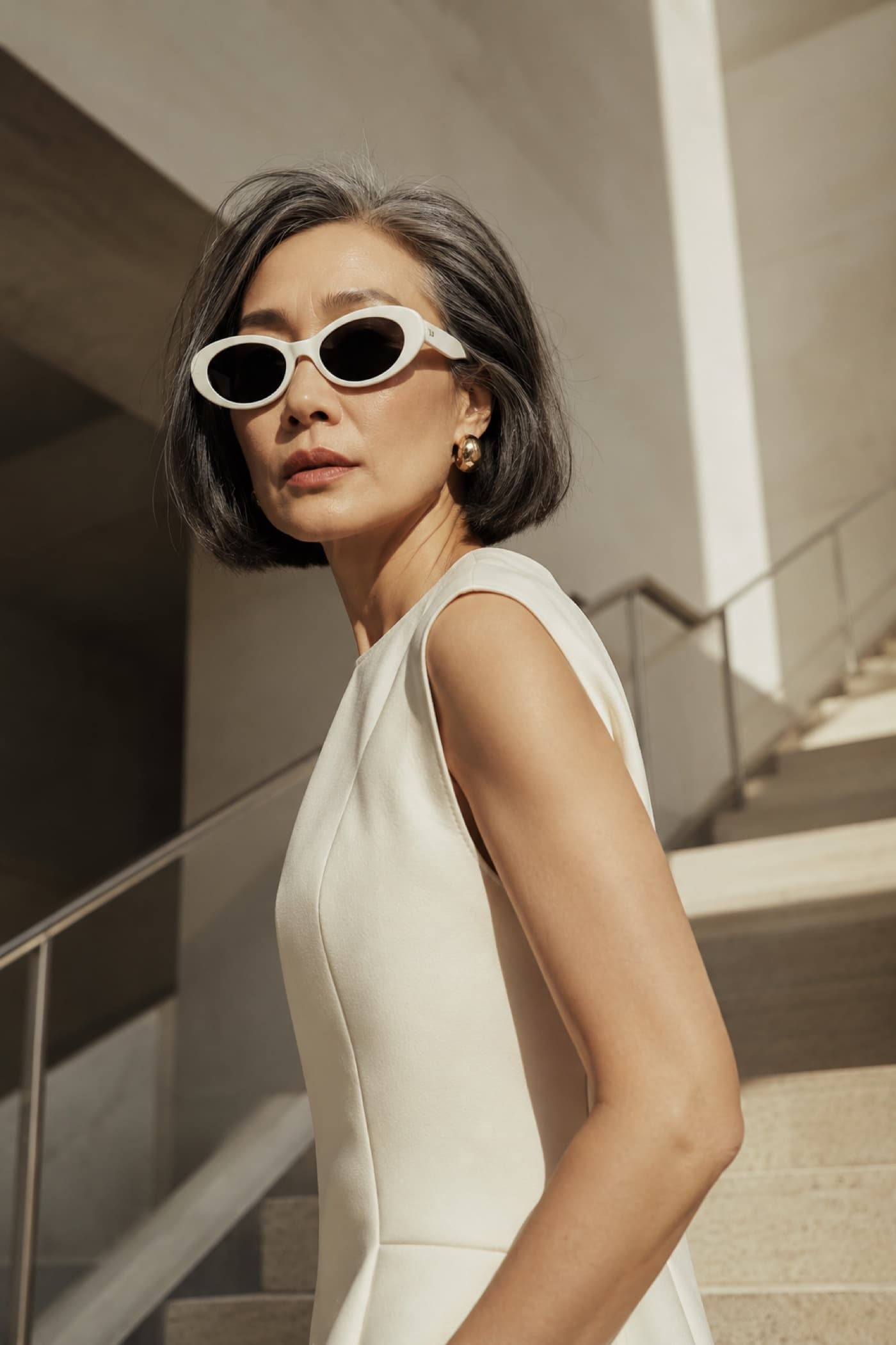 Woman with a silver bob in white oval sunglasses and a cream sleeveless dress on sunlit concrete stairs.