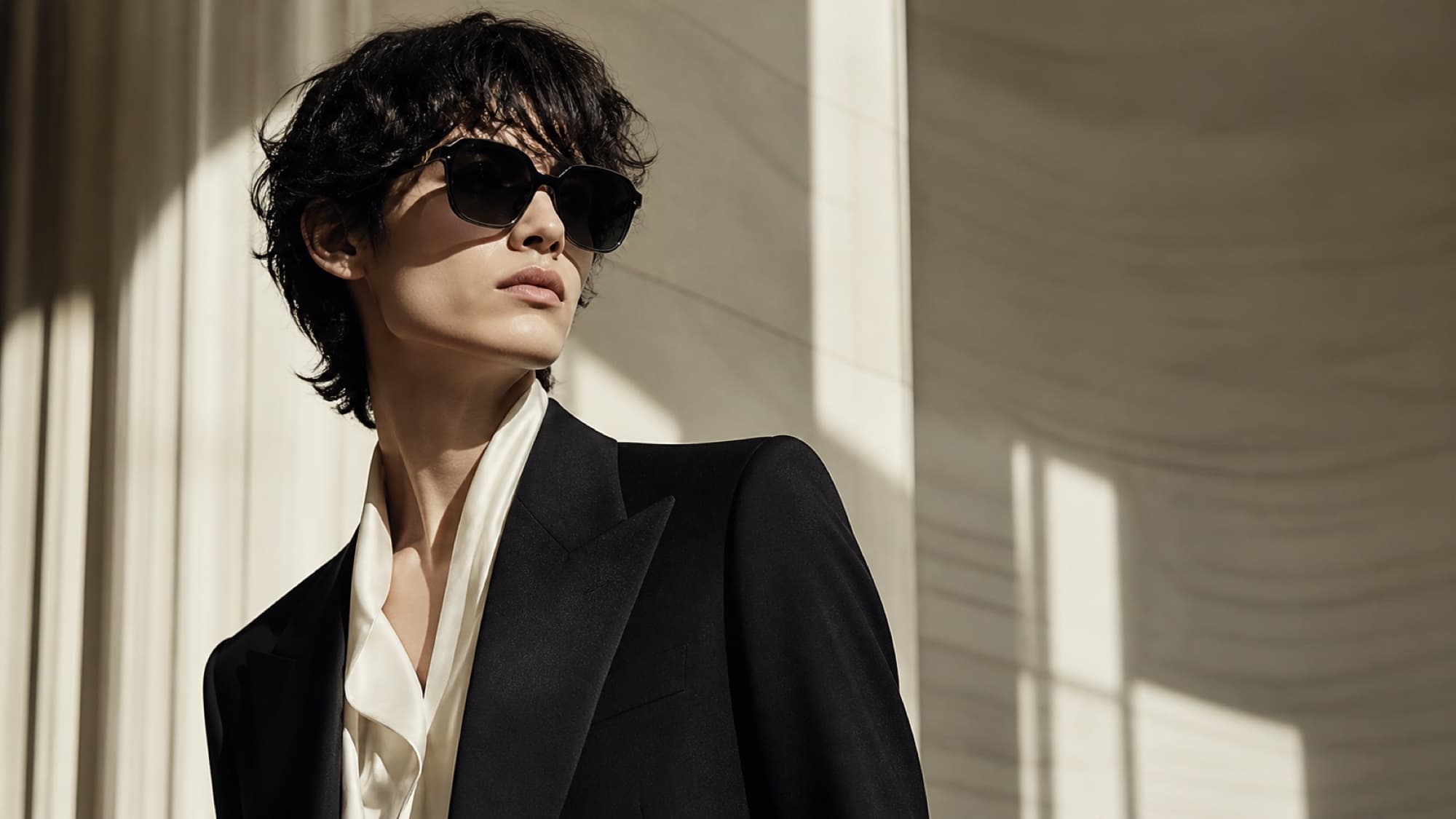 A person with tousled dark hair in a sharp black tuxedo and cream silk neck-tie blouse, photographed in dramatic side light, wearing oversized geometric sunglasses with a subtle hexagonal cut.